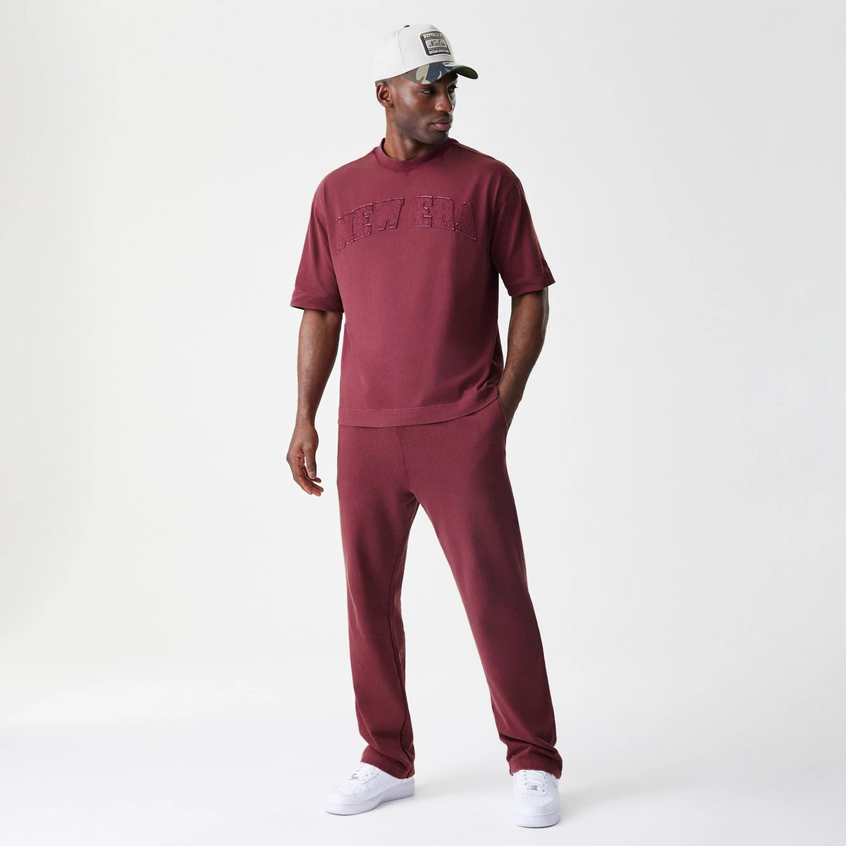 New Era Washed Dark Red Straight Leg Joggers, on-model view showing the deep red hue and relaxed fit, paired with a matching shirt featuring New Era lettering and a white cap with a front patch.