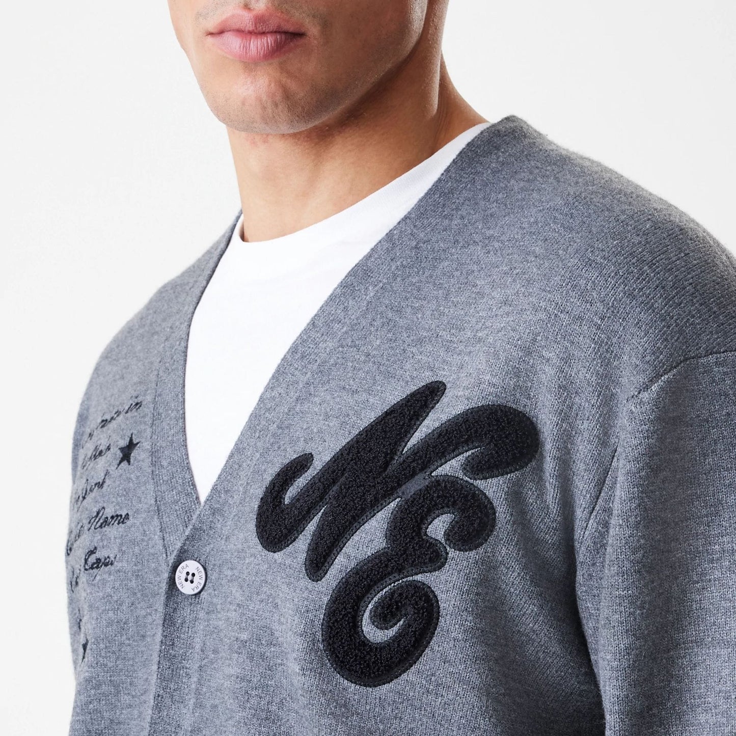New Era Dark Grey Cardigan, front view on a model showing black textured embroidered NE lettering on the left chest, contrasting button closure and a scripted design on the right side with a white T-shirt underneath.