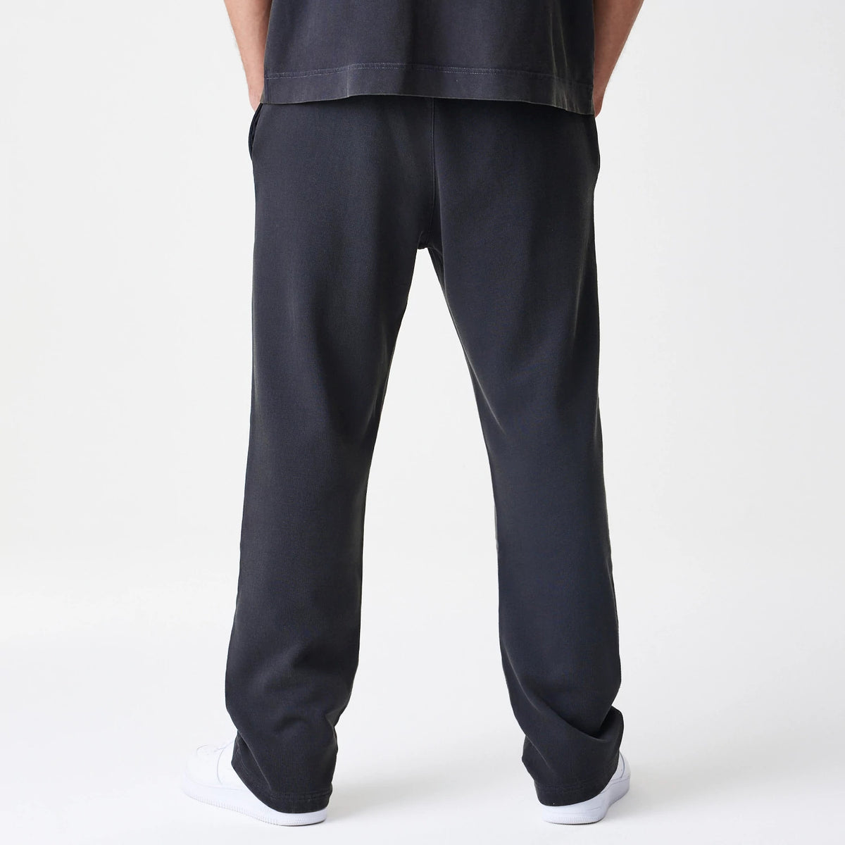New Era Washed Black Straight Leg Joggers, back view on a model showing a relaxed fit with side pockets, elastic waistband, and washed black texture, paired with white trainers for a casual look.