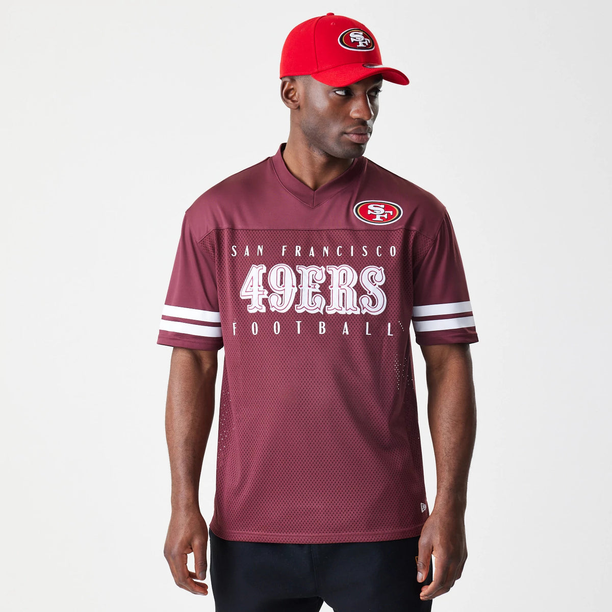 New Era San Francisco 49ers NFL Mesh Dark Red Jersey, front view on a model wearing a matching red cap, showing white printed 49ERS Football lettering and wide white stripes on each sleeve with an NFL logo on the right shoulder.