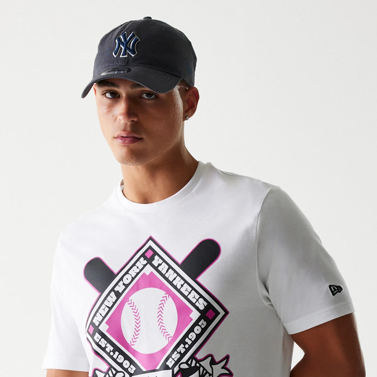 New Era New York Yankees MLB Baseball Graphic White T-Shirt, front view on-model showing a pink graphic featuring bats and baseball with New York Yankees lettering, and a small embroidered New Era logo on the left sleeve.