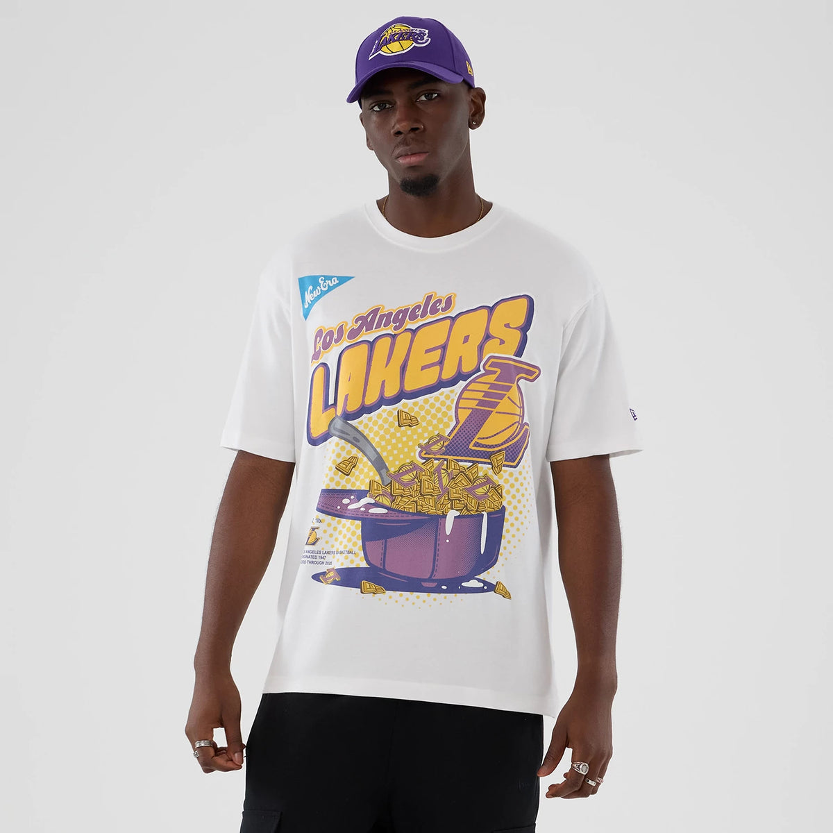 New Era LA Lakers NBA Lifestyle Graphic White T-Shirt, front view on-model showcasing a print of Los Angeles Lakers in yellow and purple, a New Era embroidered logo on the sleeve, and styled with a matching purple cap.