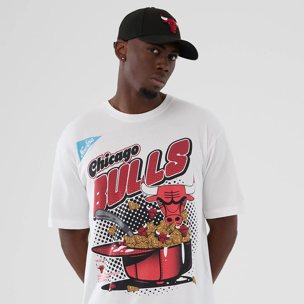 New Era Chicago Bulls NBA Lifestyle Graphic White T-Shirt, front view on-model showing a graphic featuring the Chicago Bulls logo, large red Bulls lettering, and a creative design with cereal and a ladle, worn with a black cap.