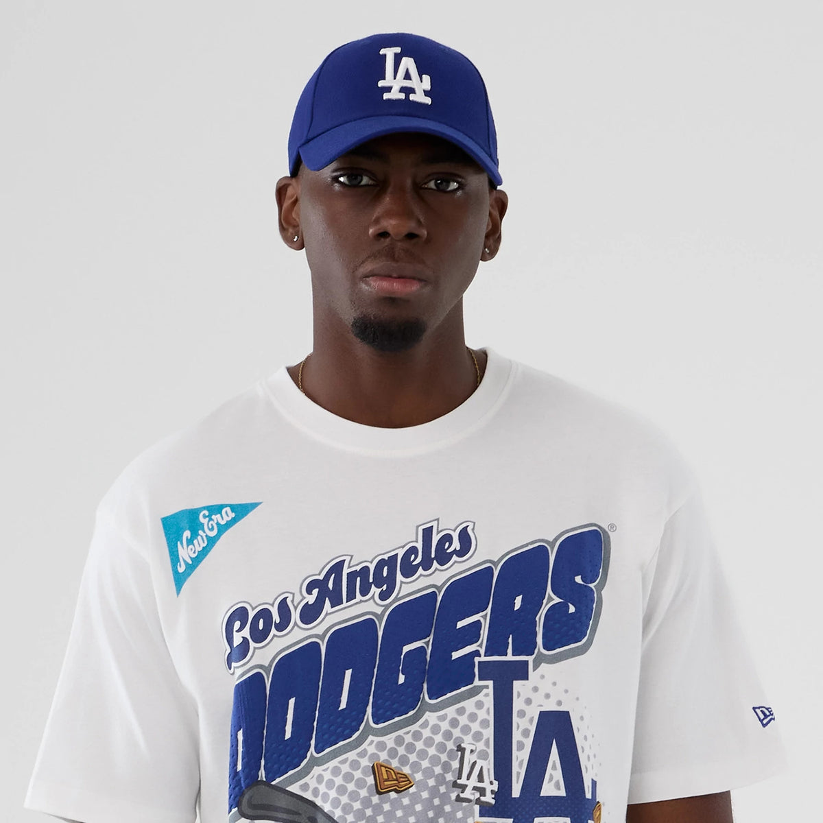 New Era LA Dodgers MLB Lifestyle Graphic White T-Shirt, front view on a model with a blue LA cap, showing a large Dodgers graphic across the chest and an embroidered New Era logo on the left sleeve.