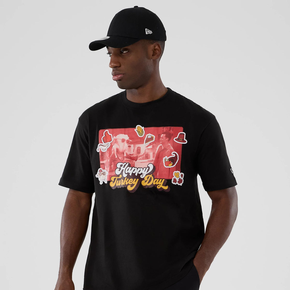 New Era Friends Turkey Head Lifestyle Graphic Black T-Shirt, front view on-model showing a red graphic with turkey head imagery and Happy Turkey Day text across the chest, worn with a plain black cap featuring an embroidered New Era logo on the side.