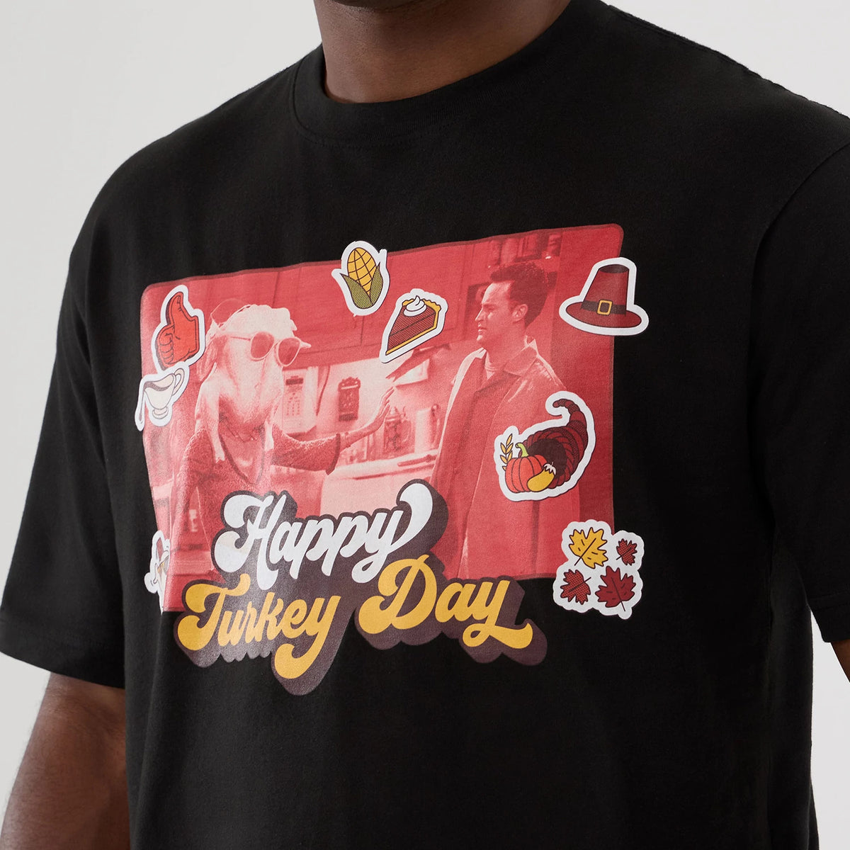 New Era Friends Turkey Head Lifestyle Graphic Black T-Shirt, front view showing a large graphic with a red tint featuring two characters and festive icons such as a corn, pie, and hat, with the words Happy Turkey Day in prominent white.
