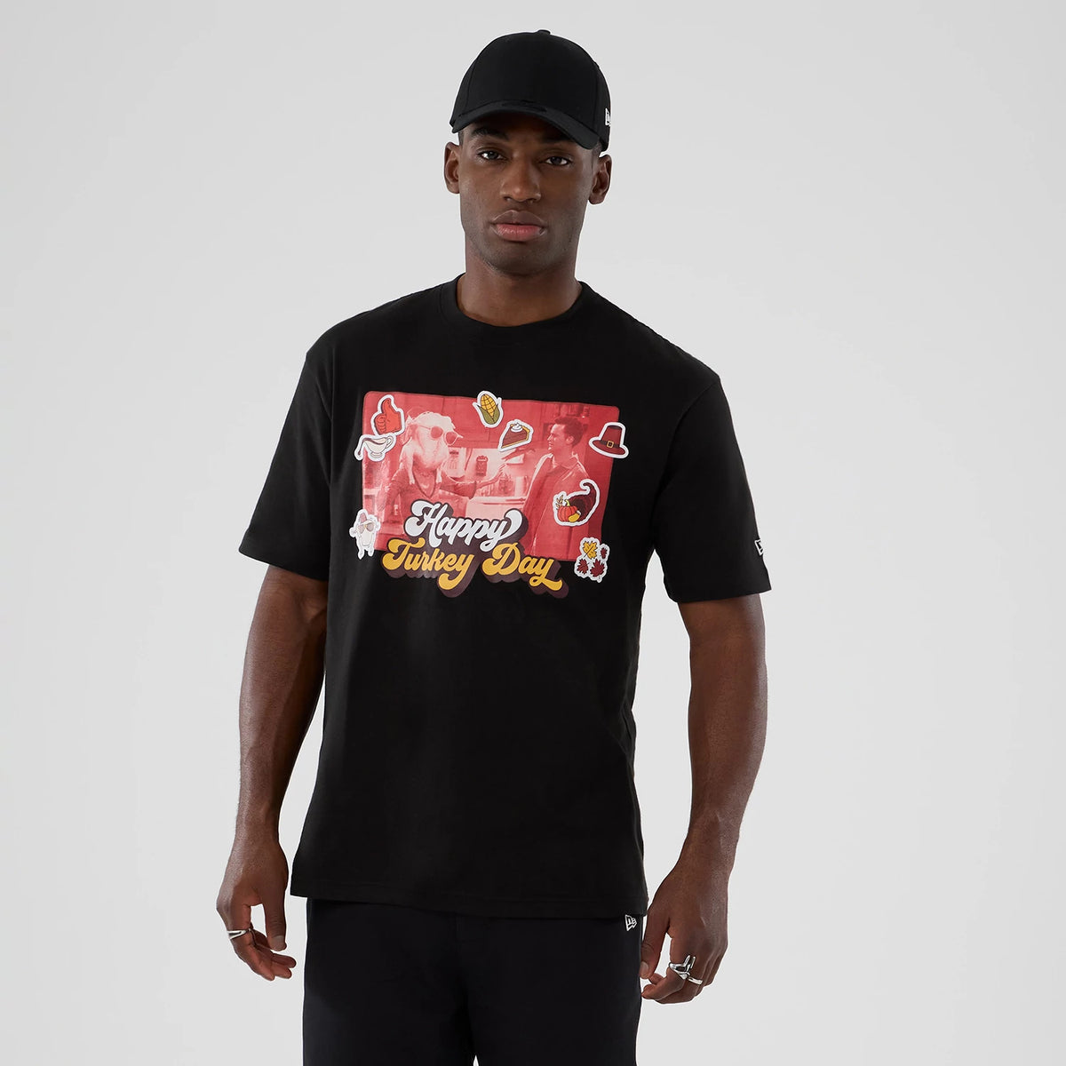 New Era Friends Turkey Head Lifestyle Graphic Black T-Shirt, front view on a model showing a red and yellow graphic featuring the words Happy Turkey Day with various small themed illustrations scattered around it.