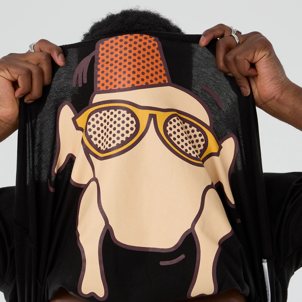 New Era Friends Turkey Head Lifestyle Graphic Black T-Shirt, front view showing a large printed graphic of a turkey with sunglasses and a red fez hat on a black background, with hands lifting the shirt to reveal more of the design.