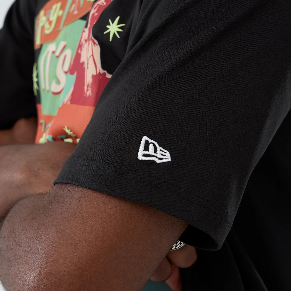 New Era Seinfeld Lifestyle Graphic Black T-Shirt, side view showing the colourful graphic print on the chest, with a white embroidered New Era logo on the sleeve of the black shirt.