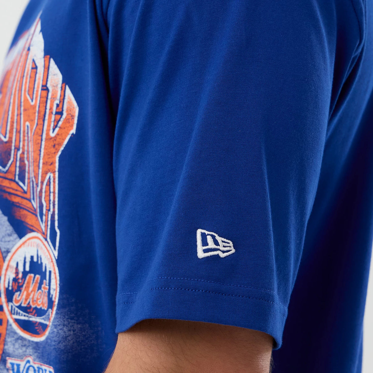 New Era New York Mets MLB Sport Classic Blue T-Shirt, side view showing the blue short sleeve with a white embroidered New Era logo near the hem and part of the New York Mets graphic with orange and white accents on the body.