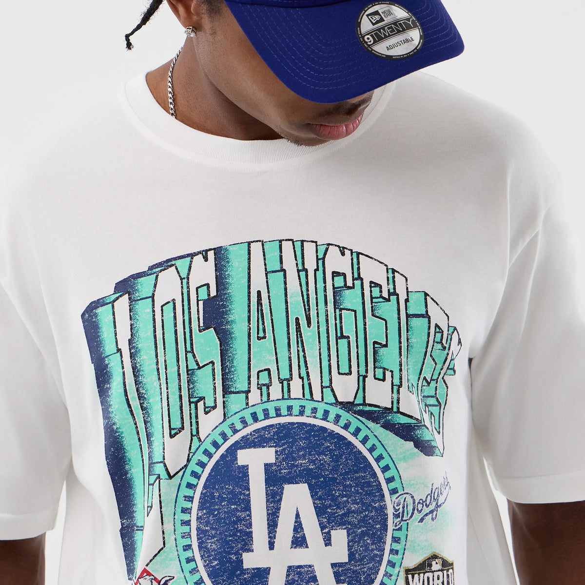 New Era LA Dodgers MLB Sport Script Classic White T-Shirt, front view on-model showing a Los Angeles graphic in blue and green with a large LA logo in the centre, worn with a matching blue cap featuring a round New Era sticker.