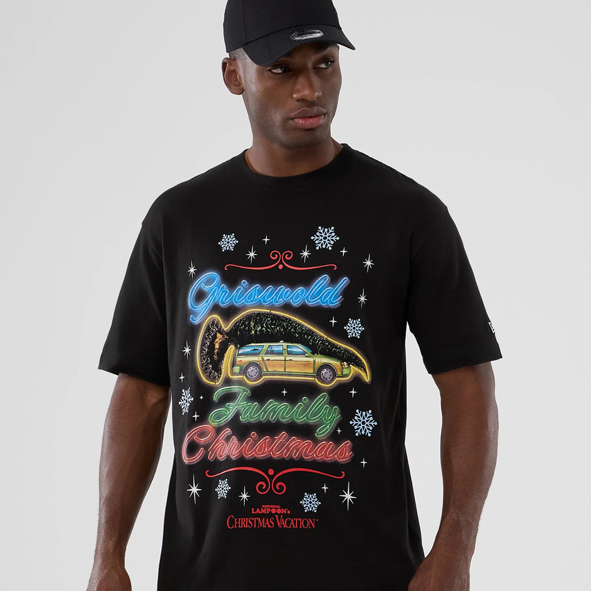 New Era National Lampoon's Christmas Vacation Lifestyle Graphic Black T-Shirt, front view on a model wearing a black cap, featuring a colourful neon-style design with Griswold Family Christmas text and a car graphic surrounded by snowflakes.
