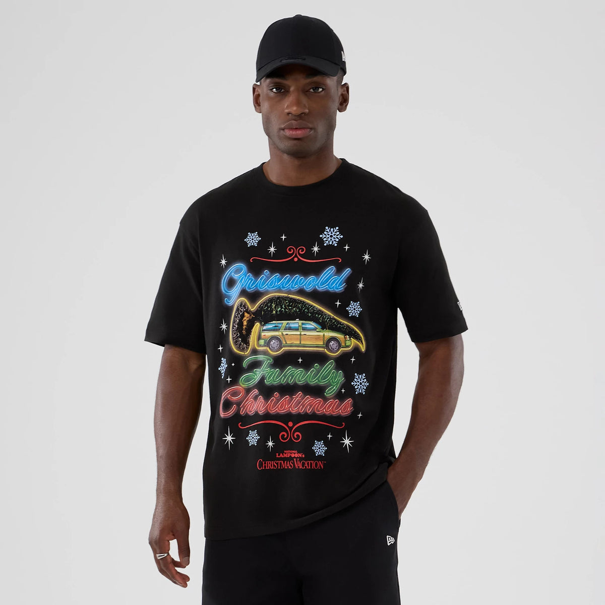 New Era National Lampoon's Christmas Vacation Lifestyle Graphic Black T-Shirt, front view showing a festive graphic with colourful lettering and a car carrying a tree, worn by a model with a black cap and embroidered New Era logo on the sleeve.