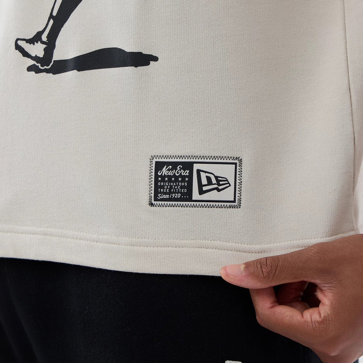 New Era New York Giants NFL Throwback Prep Light Beige T-Shirt, close-up view showing a woven New Era logo patch on the lower left side above the hem, with a black silhouette graphic at the bottom edge.