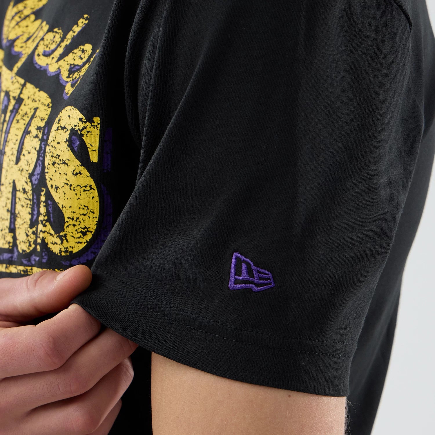 New Era LA Lakers NBA City Edition Black T-Shirt, close-up side view showing a purple embroidered New Era logo on the sleeve and the yellow and purple Lakers graphic partially on the chest.