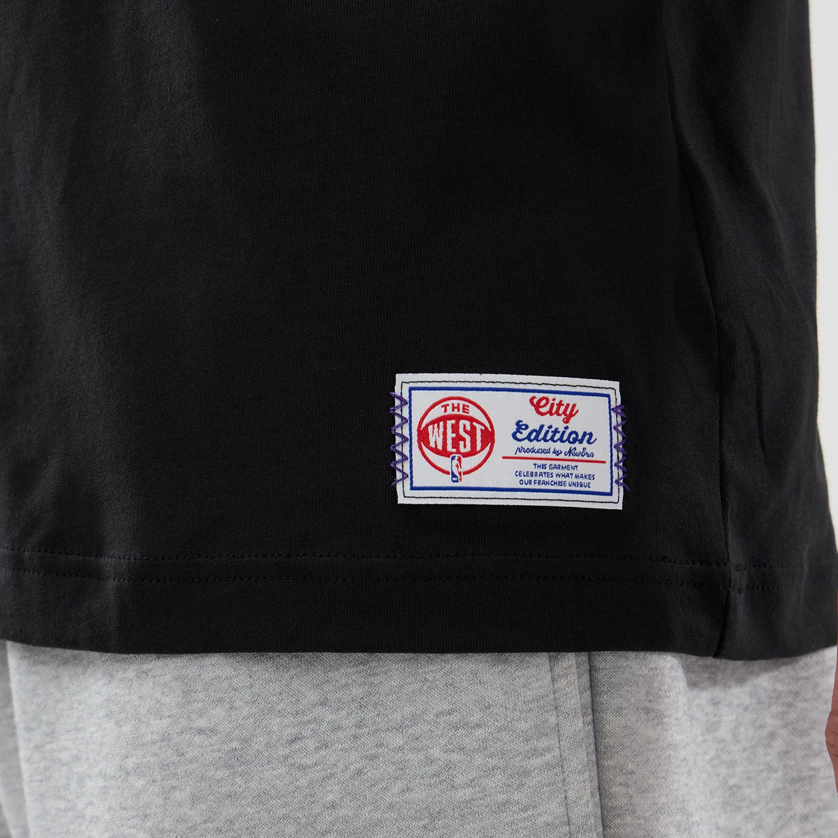 New Era LA Lakers NBA City Edition Black T-Shirt, side view showing a label near the hem with The West and City Edition in red and blue on white, contrasting against the solid black fabric of the T-shirt.