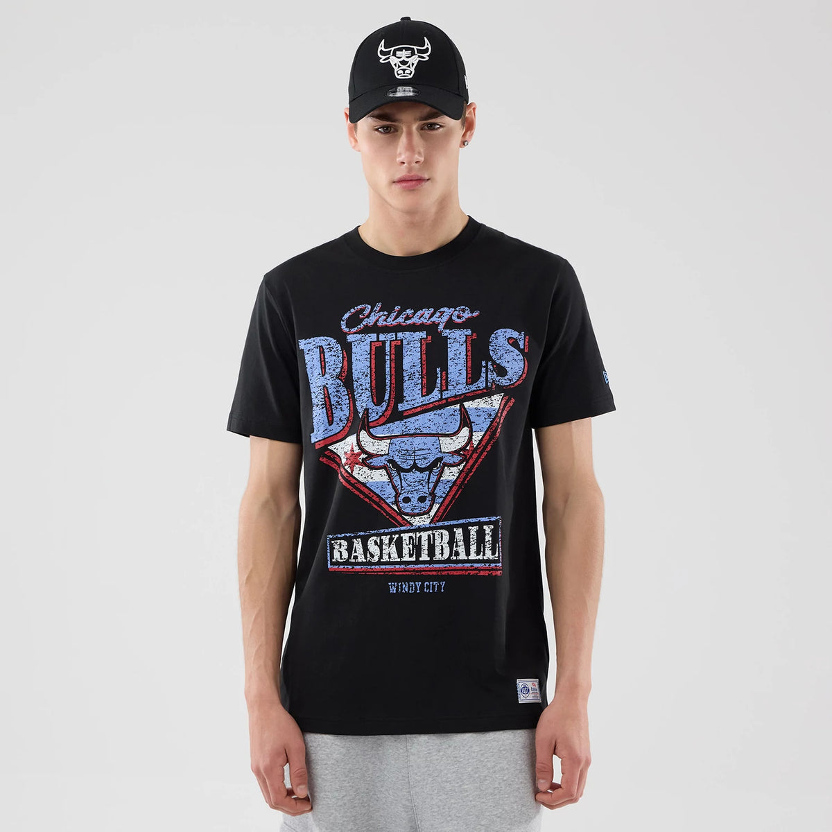 New Era Chicago Bulls NBA City Edition Black T-Shirt, front view on-model showcasing a large distressed blue and red Bulls logo with Basketball and Windy City text, paired with a black cap featuring an embroidered Bulls logo.