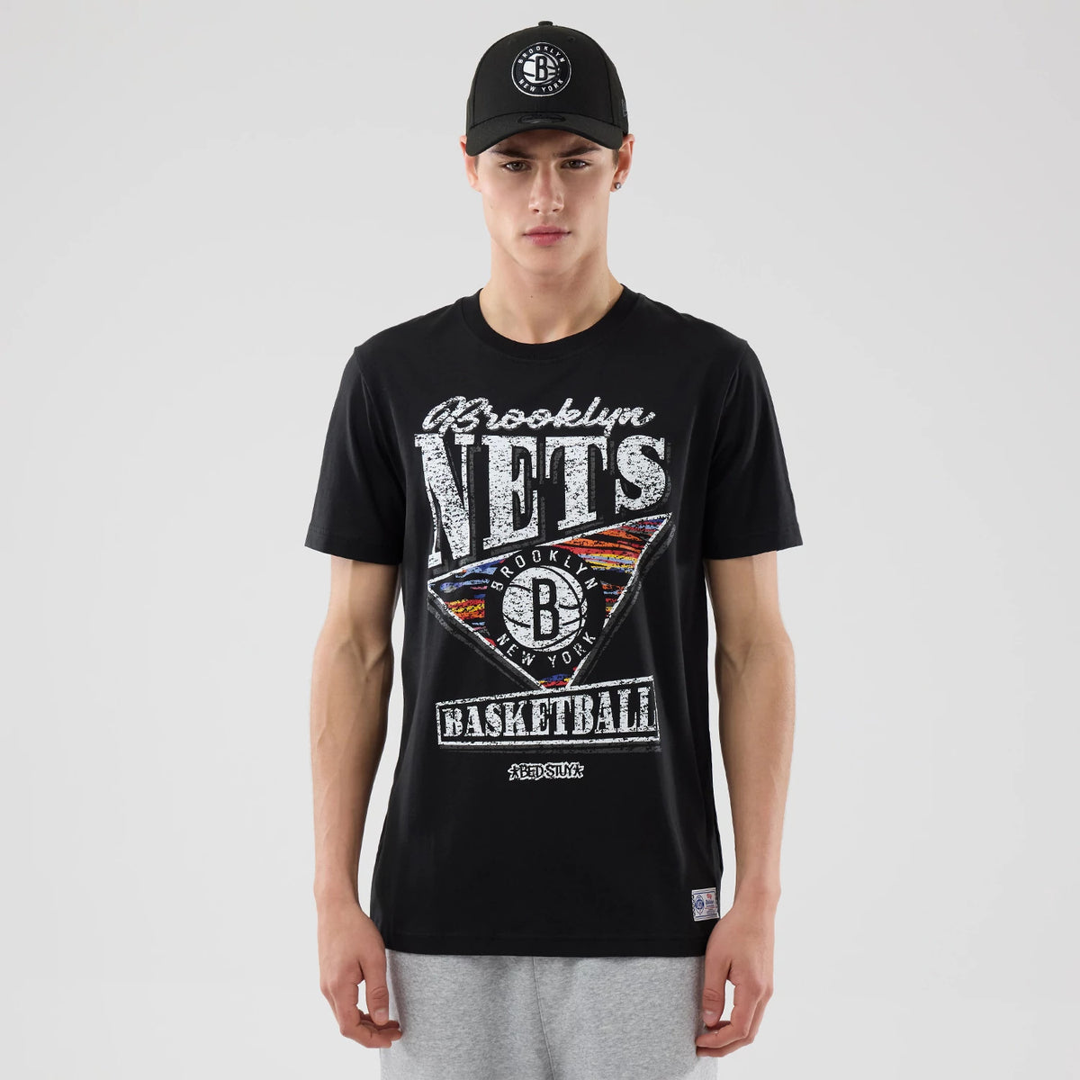 New Era Brooklyn Nets NBA City Edition Black T-Shirt, front view on-model displaying a distressed graphic with Brooklyn Nets and Basketball text, a triangular emblem with a black circle logo in the centre, styled with a matching cap.