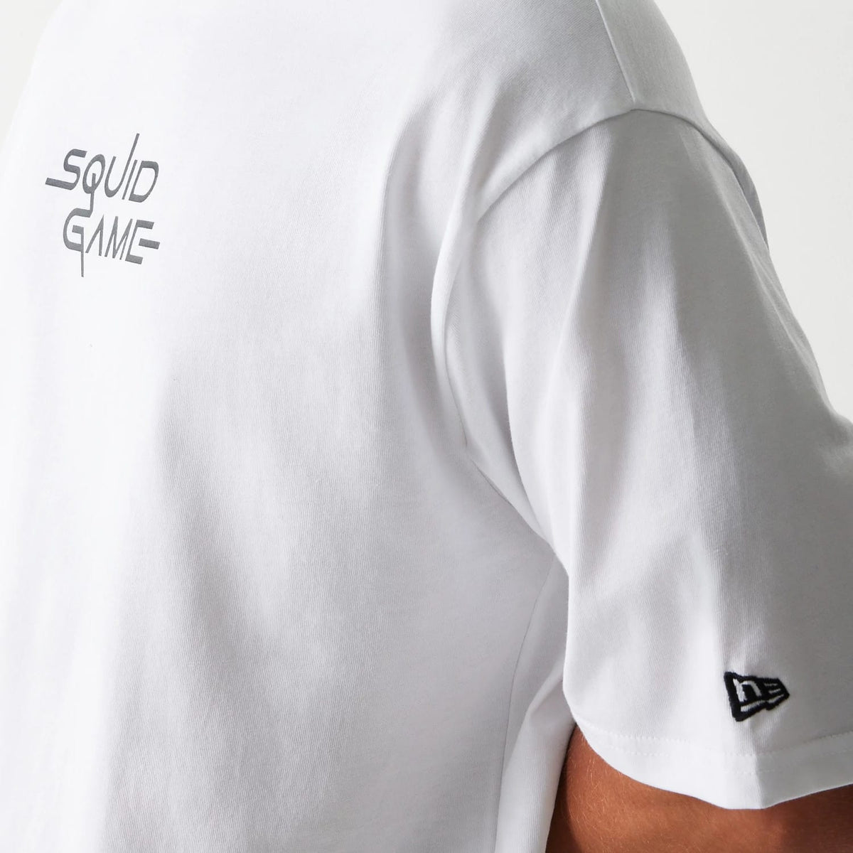 New Era Netflix Squid Game White Oversized T-Shirt, side view showing the black embroidered New Era logo on the sleeve and the grey Squid Game text graphic on the upper back, set against the plain white fabric.