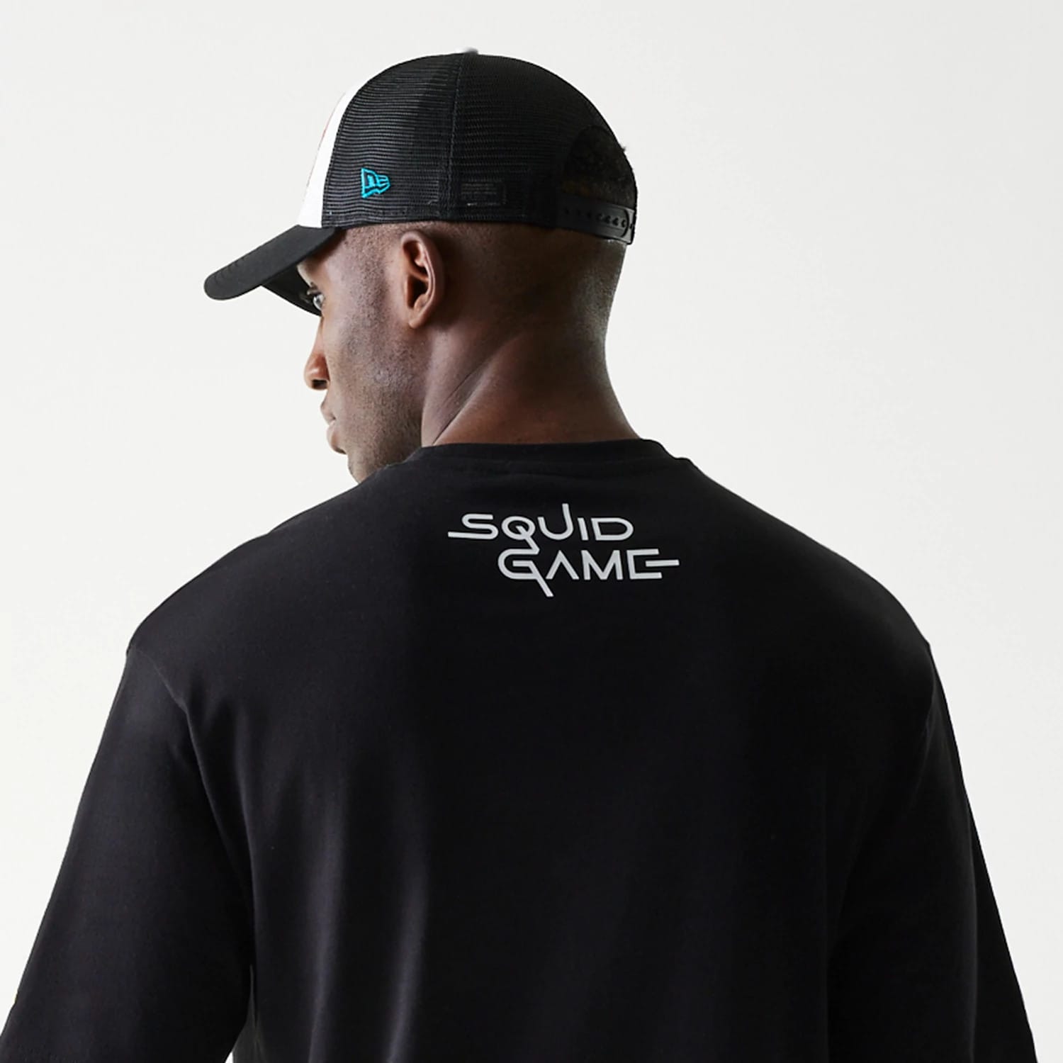 New Era Netflix Squid Game Black Oversized T-Shirt, back view showing the white printed Squid Game logo across the upper back, with a model styled wearing a black and white cap that features a blue embroidered New Era logo on the side.