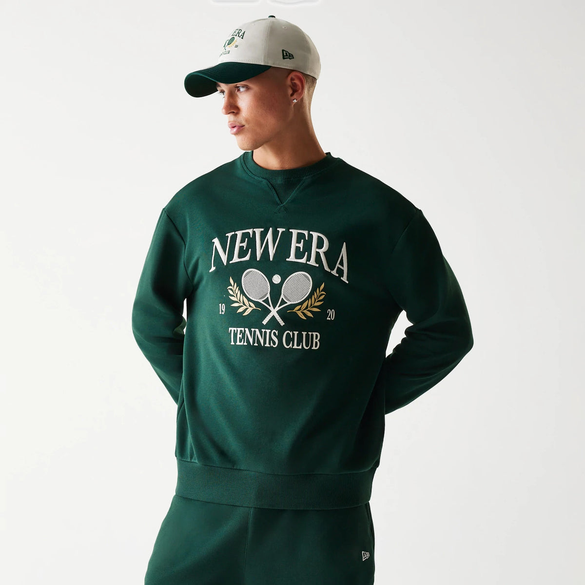 New Era Dark Green Oversized Crew Neck Sweater, front view on a model with matching dark green and white cap, featuring large white New Era Tennis Club embroidery with crossed rackets and laurel graphics on the chest.