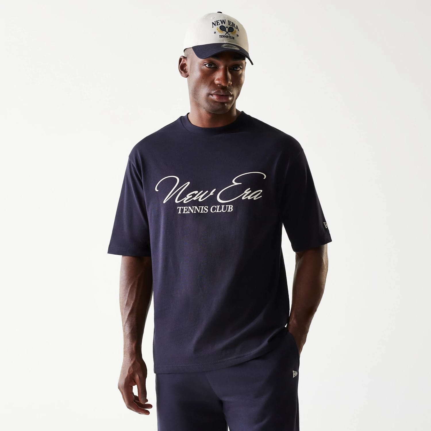 New Era Navy Oversized T-Shirt, front view on a model featuring large white New Era Tennis Club script across the chest and a small embroidered New Era logo on the left sleeve, styled with matching navy trousers and a cream cap.