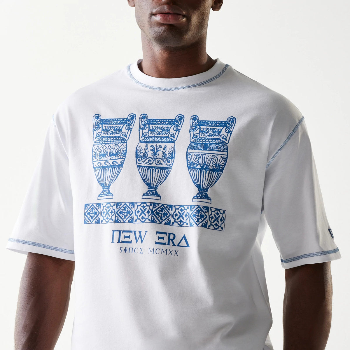 New Era Orthodox Vase White Oversized T-Shirt, front view on a model showing three intricate blue vase graphics and a patterned strip above the printed New Era logo with Greek script at the lower front.