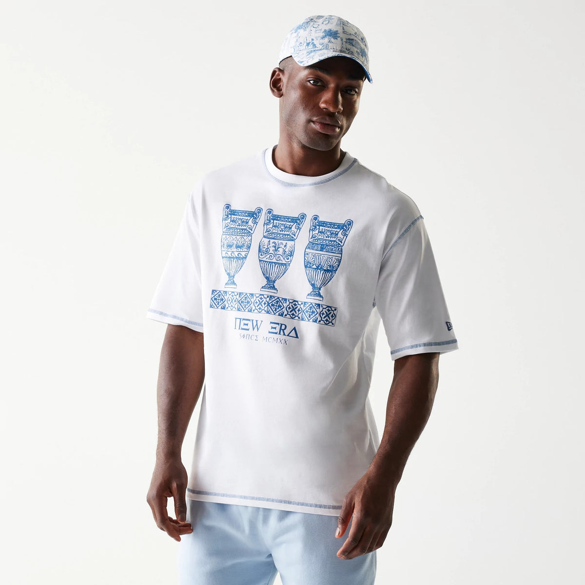 New Era Orthodox Vase White Oversized T-Shirt, front view on-model showing three detailed blue vases printed across the chest with a New Era logo beneath, blue trims on the sleeves and collar, paired with a patterned cap.