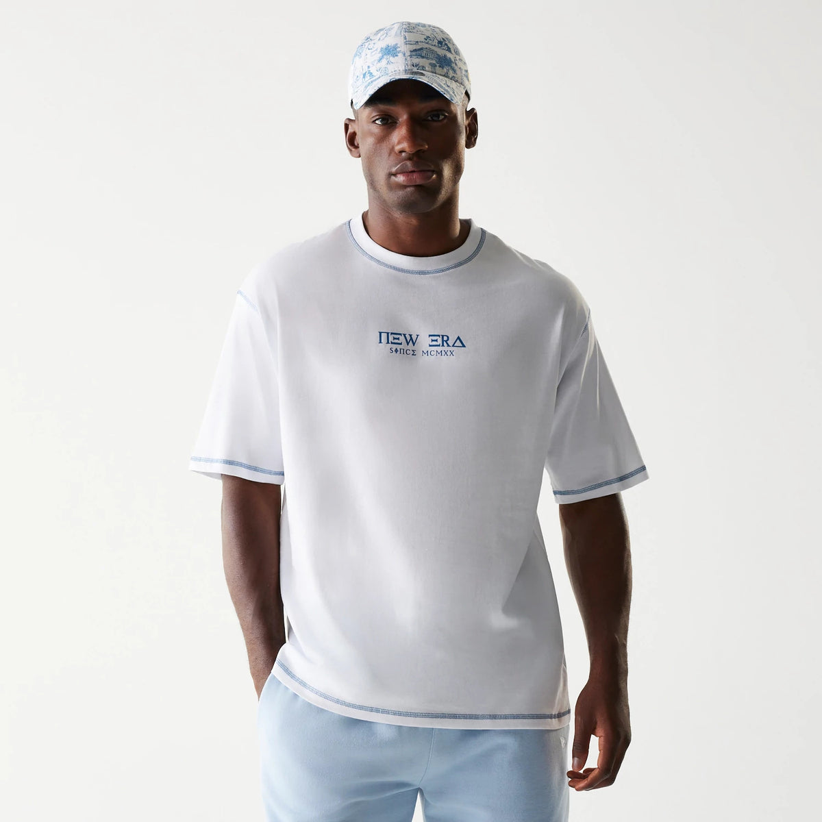 New Era Orthodox Church White Oversized T-Shirt, front view on-model showing a large New Era embroidered logo with added text in blue on the chest, complemented by light blue trim on the sleeves and collar.