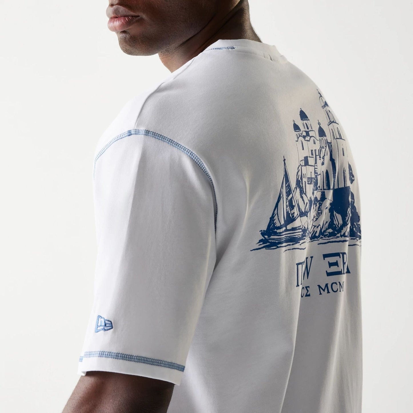 New Era Orthodox Church White Oversized T-Shirt, three-quarter back view showing intricate blue graphic of a church on the upper back, blue printed New Era logo on the left sleeve and blue stitching along the shoulder seams.