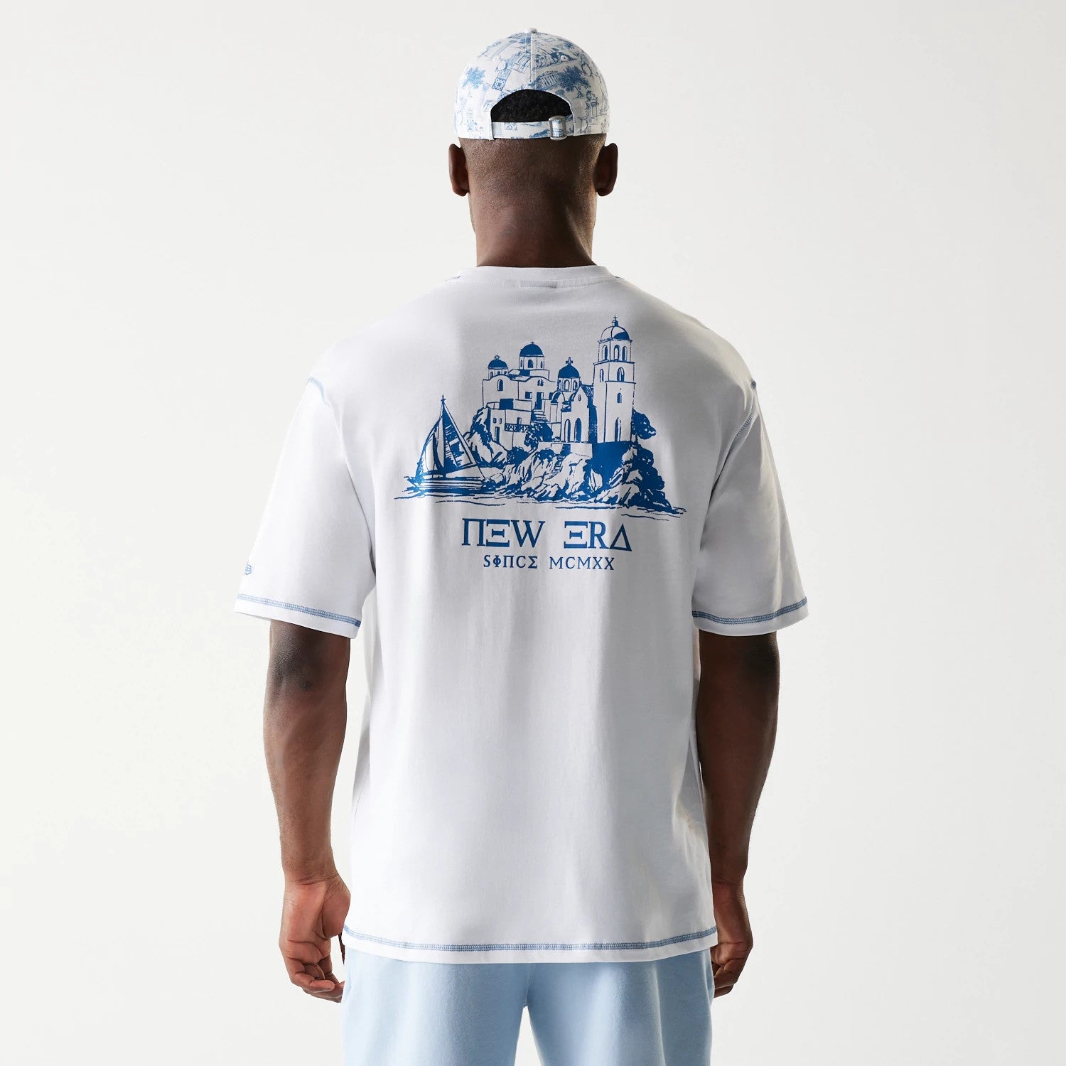 New Era Orthodox Church White Oversized T-Shirt, back view on-model featuring a detailed blue graphic of an Orthodox church and boat on the upper back, New Era lettering and since MCMXX printed beneath, with blue trim on sleeves and hem.