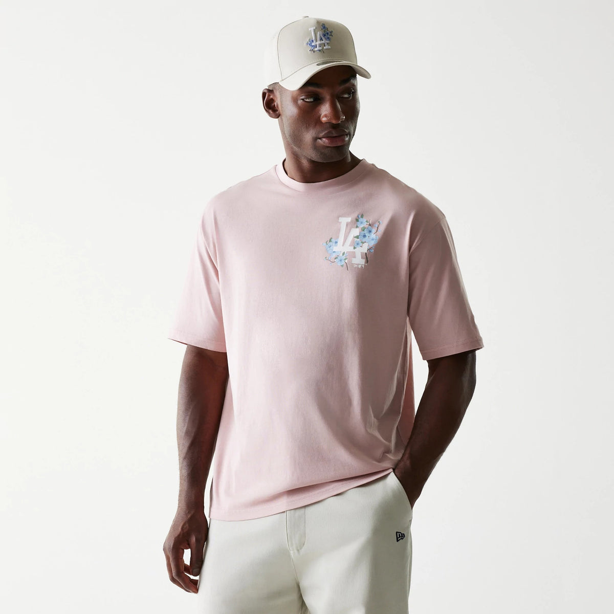 New Era LA Dodgers MLB Floral Pastel Pink Oversized T-Shirt, front view on a model with a white cap, featuring an embroidered LA logo surrounded by floral graphics on the left chest area of the pastel pink fabric.