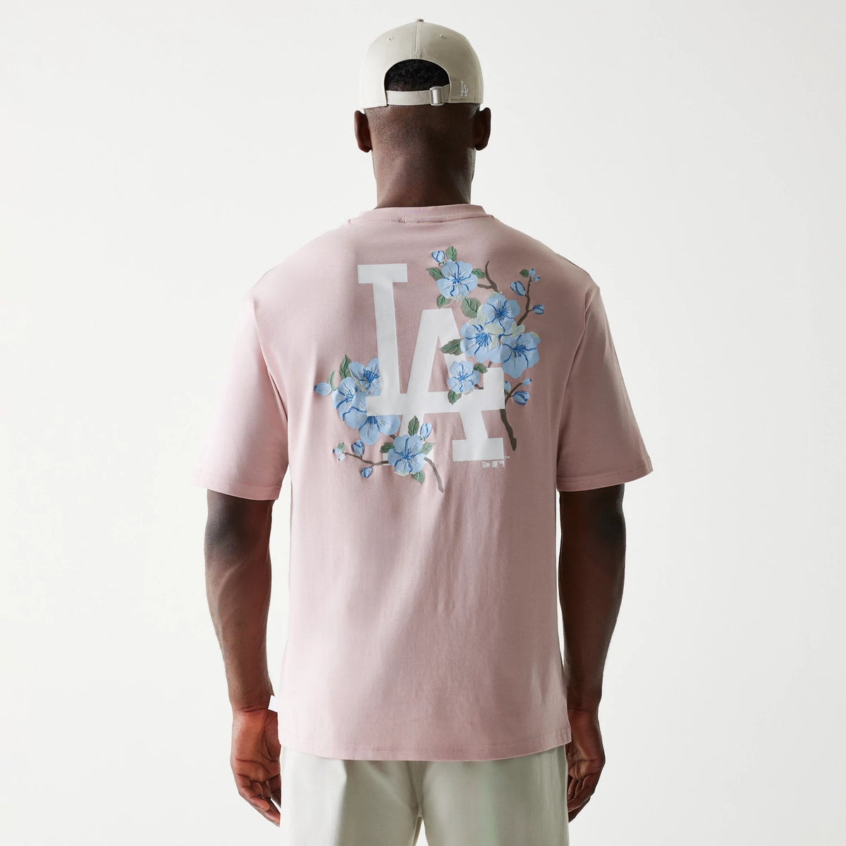 New Era LA Dodgers MLB Floral Pastel Pink Oversized T-Shirt, back view showing a large white LA graphic with pastel blue floral embellishments across the upper back, styled on-model with a complementary cap.