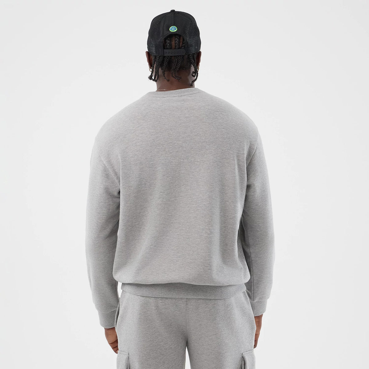 New Era Sakamoto Days Netflix Grey Crew Neck Sweater, back view on a model showing the plain grey fabric with ribbed trim at the waist and cuffs, paired with matching grey trousers and a black mesh cap.