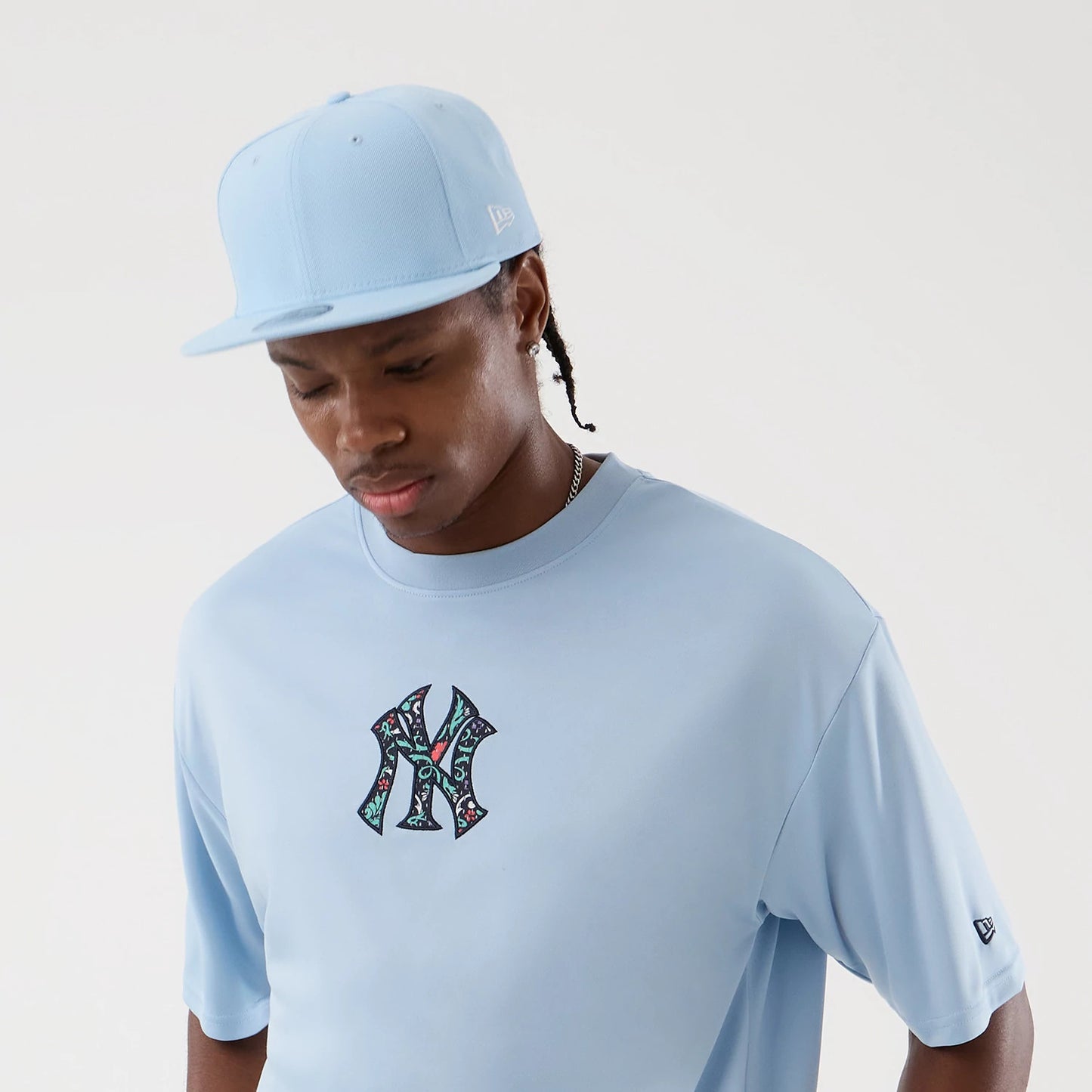 New Era New York Yankees MLB Mesh Infill Pastel Blue Oversized T-Shirt, front view on-model showing large floral-patterned NY logo centred on the chest and a small embroidered New Era logo on the left sleeve, paired with a pastel blue cap.