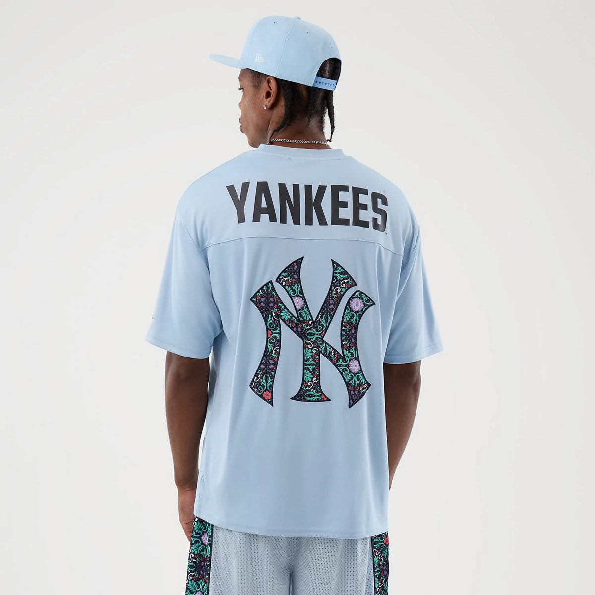 New Era New York Yankees MLB Mesh Infill Pastel Blue Oversized T-Shirt, back view on a model with a pastel blue cap, featuring Yankees lettering across the shoulders and a large NY logo with a colourful patterned infill on the centre back.
