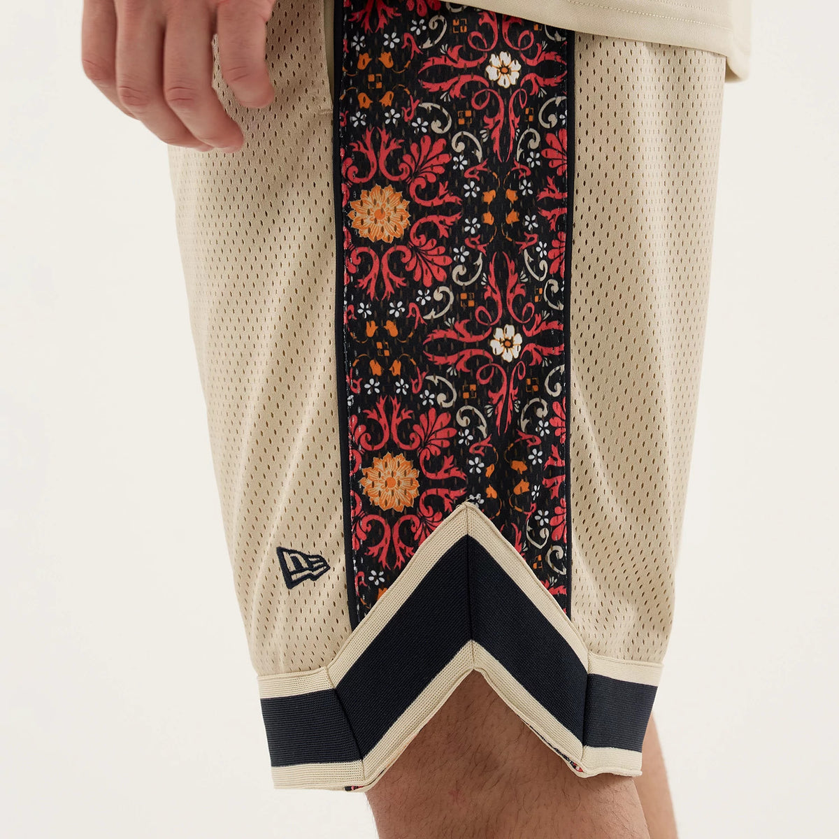 New Era Mesh Light Beige Mesh Shorts, side view showing a detailed patterned strip in red, orange, and white down the leg, a black embroidered New Era logo near the hem, and navy trims at the edge.