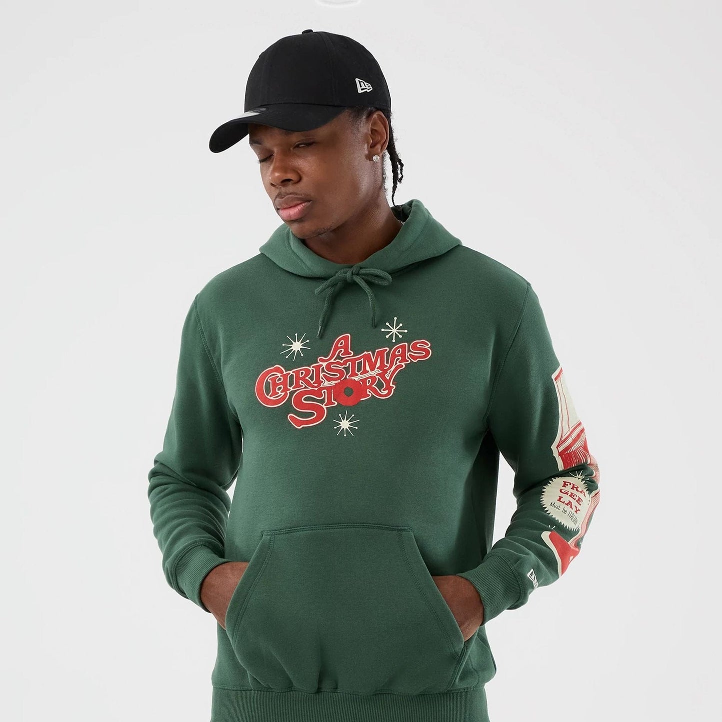 New Era A Christmas Story Major Award Dark Green Pullover Hoodie, front view on-model showing red A Christmas Story lettering and star graphics centred on the chest, with a printed leg lamp and text detail on the left sleeve.