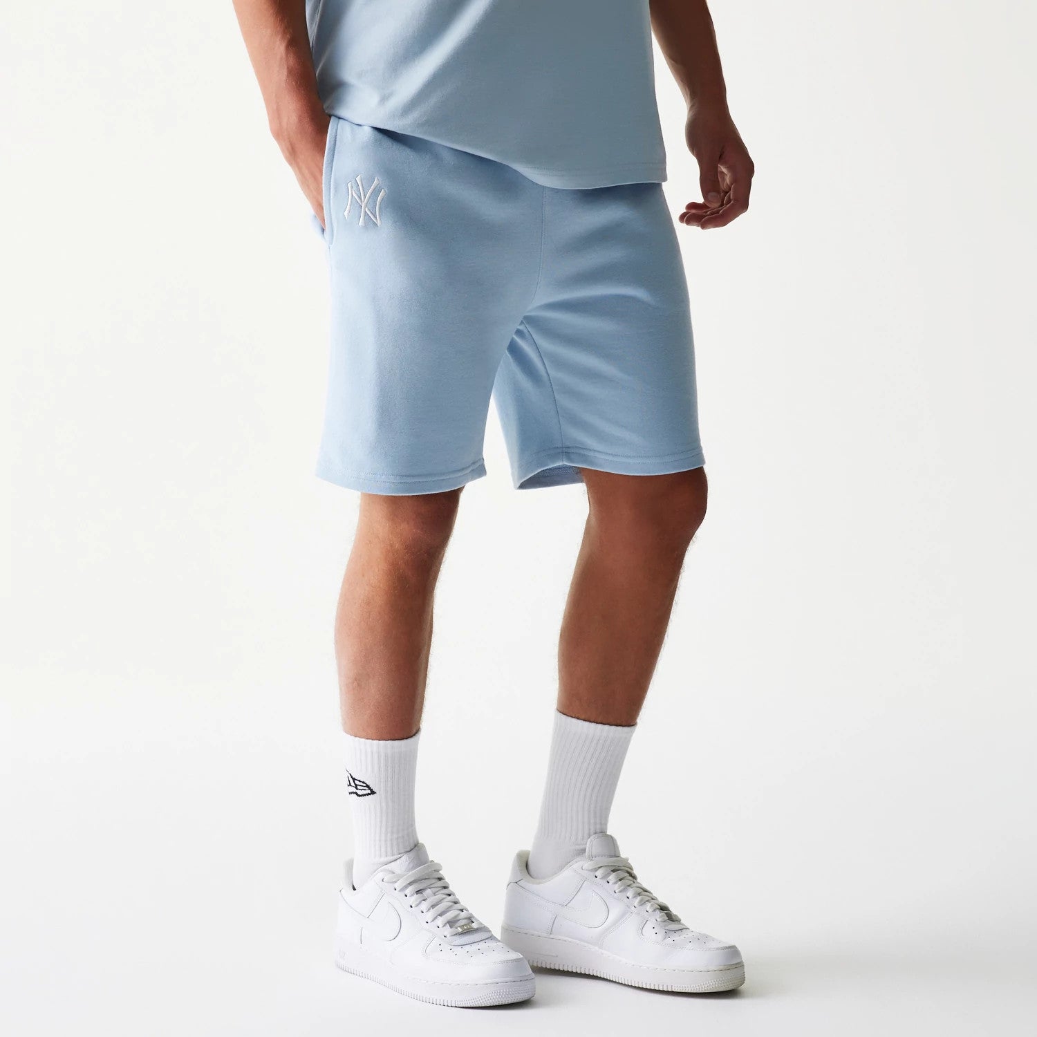 New Era New York Yankees MLB League Essential Midi Pastel Blue Shorts, front three-quarter view on a model showing the pastel blue fabric with an embroidered white New York Yankees logo on the left leg, paired with white trainers and socks.