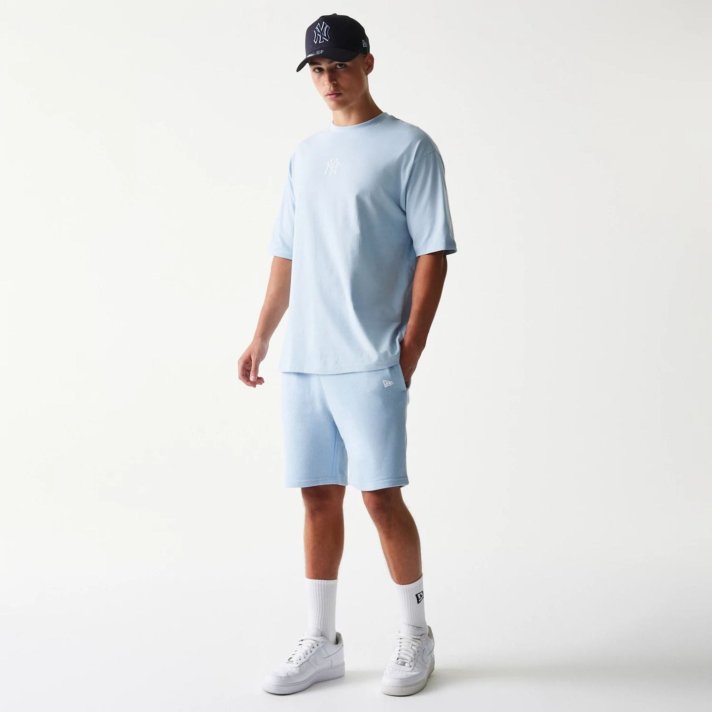 New Era New York Yankees MLB League Essential Midi Pastel Blue Shorts, on-model view with a matching pastel blue top, displaying an embroidered New Era logo on the left thigh and paired with white socks and trainers.
