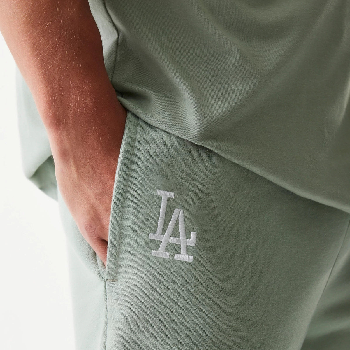 New Era LA Dodgers MLB League Essential Midi Pastel Green Shorts, side view highlighting an embroidered white LA logo on the left pocket, with the shorts styled casually on a model demonstrating relaxed fit and comfort.
