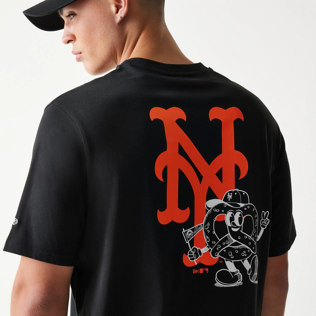 New Era New York Mets MLB Food Graphic Black T-Shirt, back view showing a large orange Mets logo with a playful pretzel character graphic below, on a model styled with a black cap.
