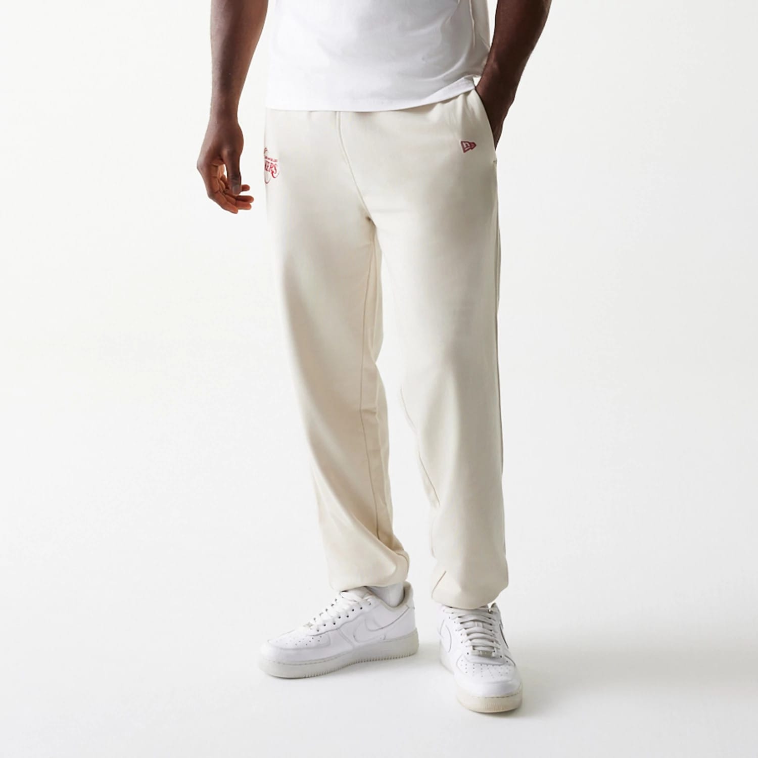 New Era LA Lakers NBA League Essential Cream Joggers, front view on a model showing LA Lakers logo embroidered in red on the right thigh, with pockets and a relaxed fit, paired with white trainers and a plain white T-shirt.