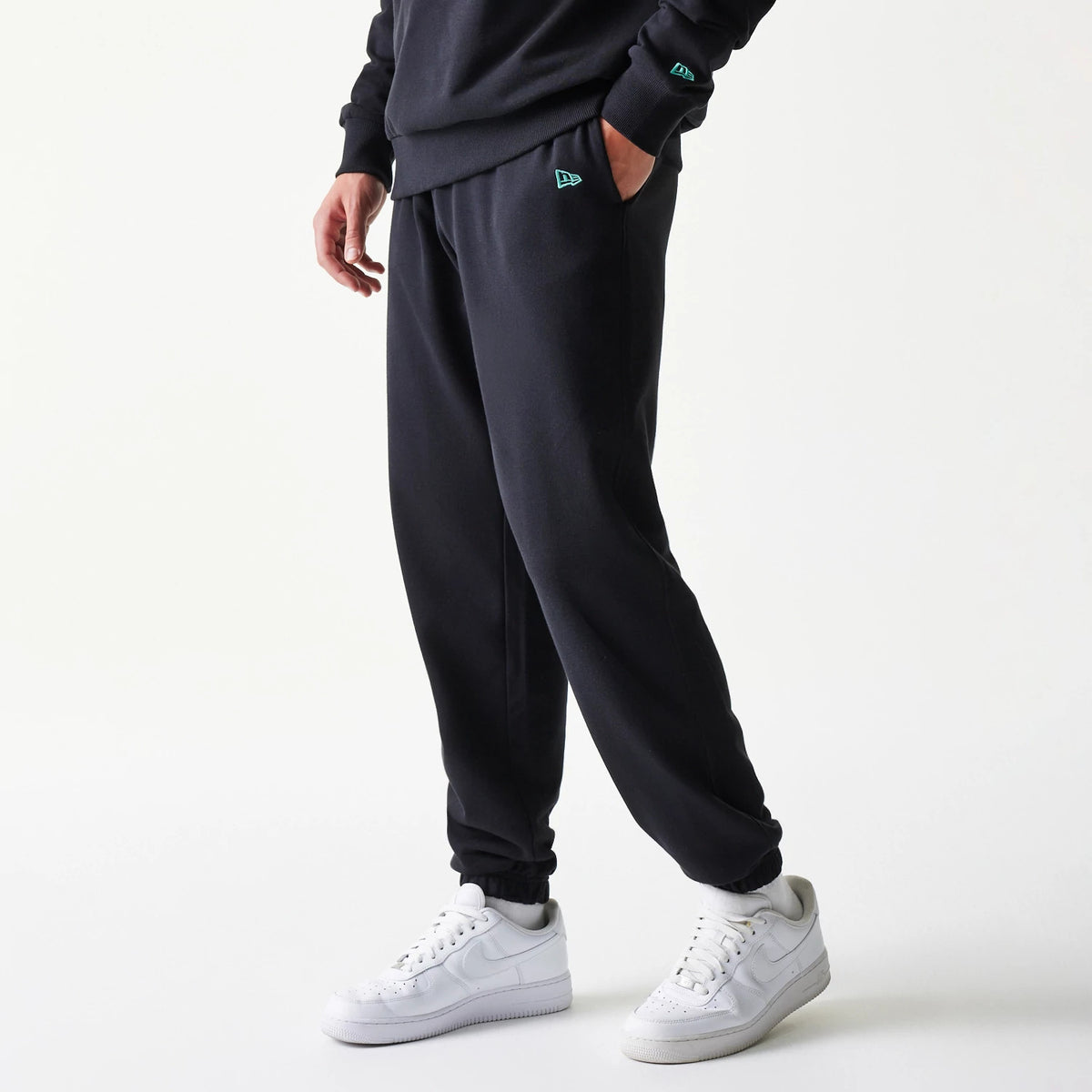 New Era Chicago Bulls NBA League Essential Black Joggers, side view on a model showing relaxed fit with teal New Era flag logo on the left pocket and cuff, paired with white trainers for a casual look.