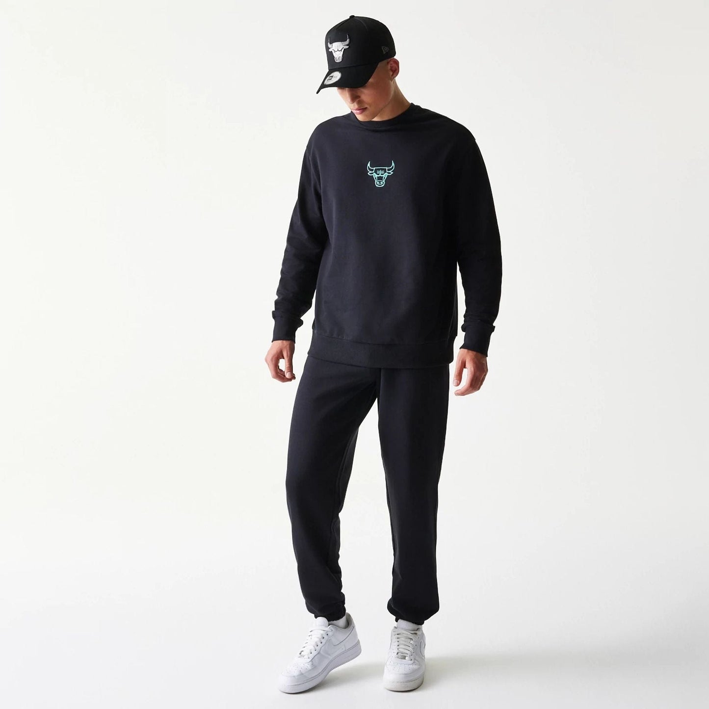 New Era Chicago Bulls NBA League Essential Black Joggers, on a model with matching black sweatshirt and cap, featuring a relaxed fit and elasticated cuffs, styled with white trainers for a casual look.