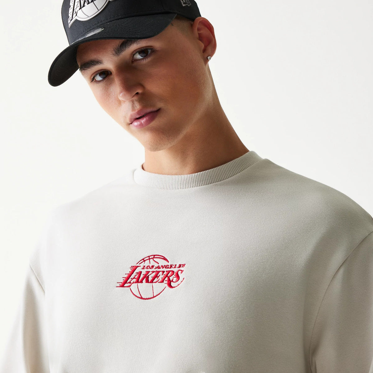 New Era LA Lakers NBA Cream Oversized Crew Neck Sweater, front view on a model wearing a black cap, featuring the red embroidered Lakers logo on the chest and showcasing the ribbed crew neckline.