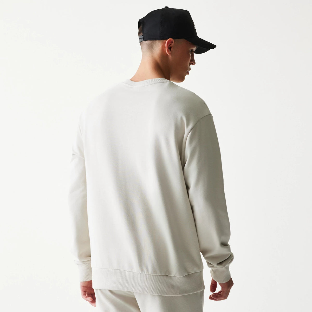 New Era LA Lakers NBA Cream Oversized Crew Neck Sweater, back view on a model paired with a black cap, showing the simple cream colour and relaxed fit with ribbed cuffs and hem for a casual look.