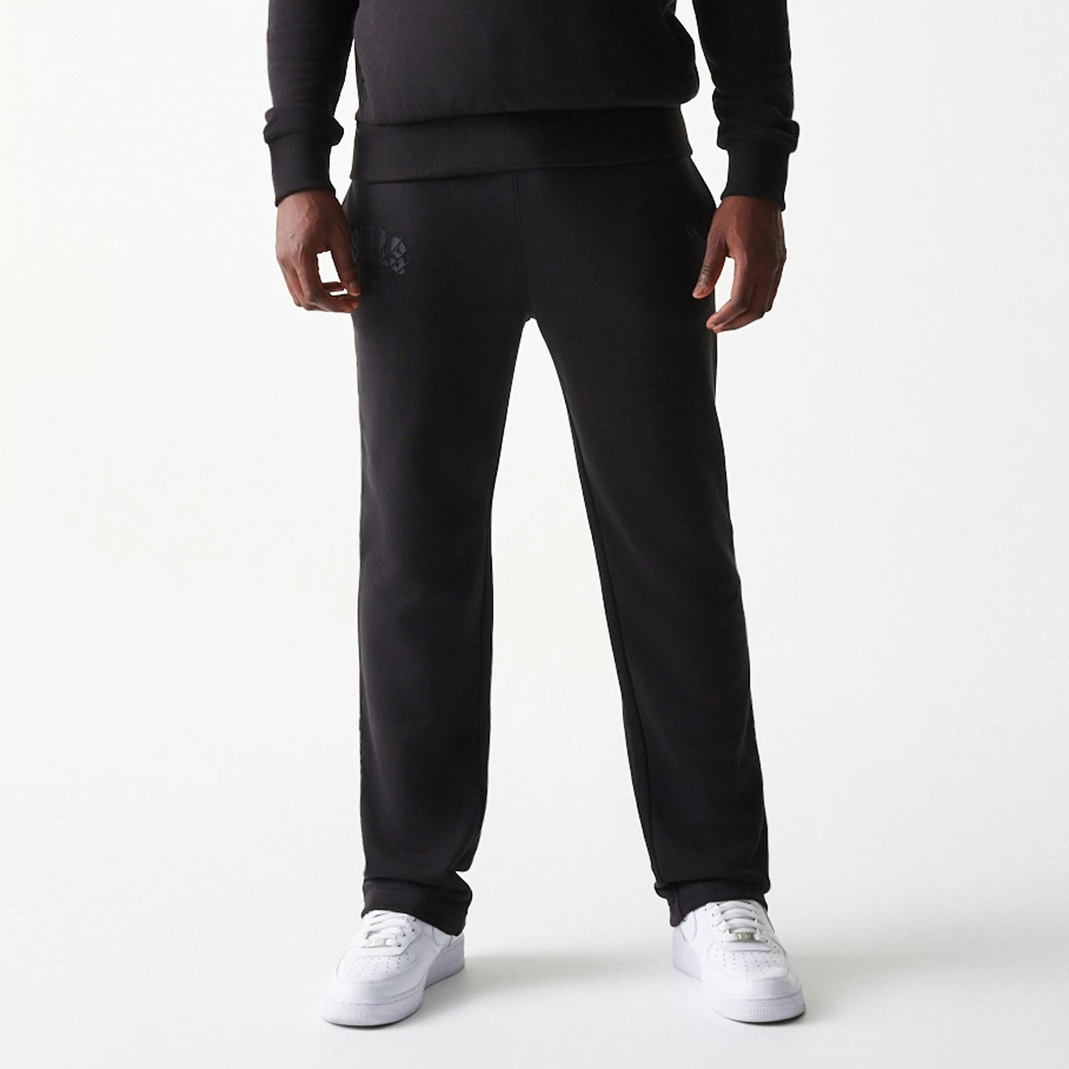 New Era Chicago Bulls NBA Black Straight Leg Joggers, front view on a model wearing white trainers, featuring the Chicago Bulls logo printed on the upper left leg, and a relaxed fit design with ribbed ankle cuffs.