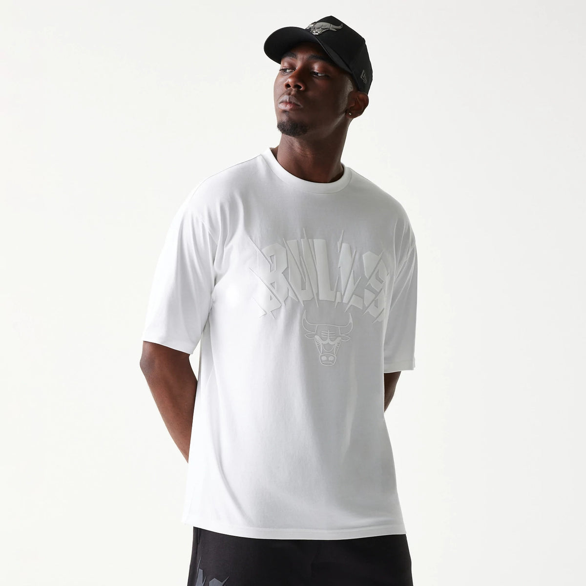 New Era Chicago Bulls NBA White Oversized T-Shirt, front view on a model displaying the raised BULLS lettering across the chest with the bull logo underneath, styled with a black cap featuring an embroidered logo.