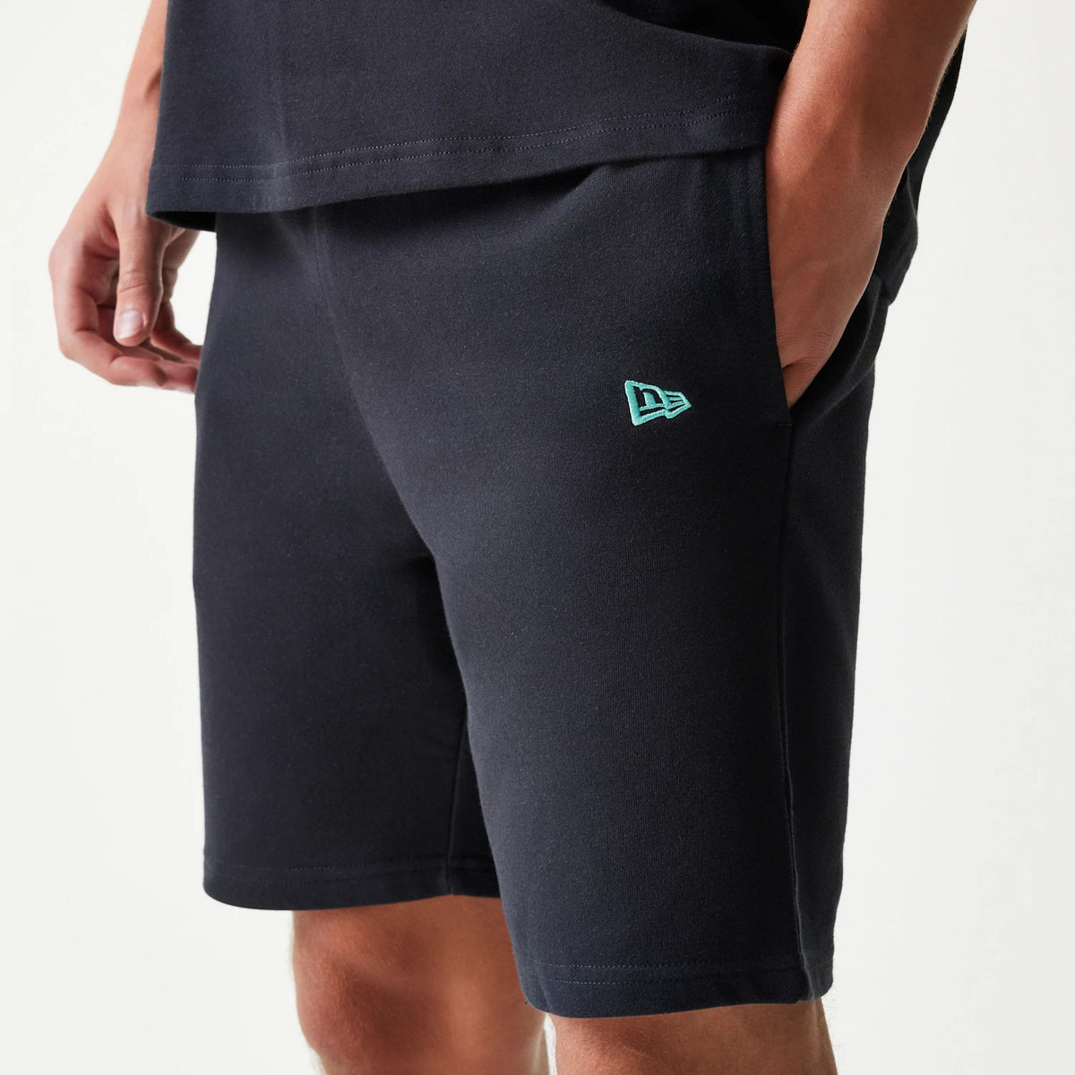 New Era Chicago Bulls NBA League Essential Black Shorts, side view showing a turquoise embroidered New Era logo on the lower left leg, worn by a model with hands in pockets highlighting a relaxed fit and plain black design.